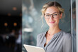 © Lady O - A smiling business woman holding a tablet and standing near a window in an office.
