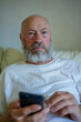 © Liana - Senior bearded man using smartphone while sitting at home