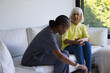© WavebreakMediaMicro - Diverse carer in scrubs leaning to senior woman in yellow-blouse on sofa in living-room smartwatch