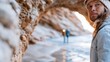 © Pinklife - A young man with long hair in winter attire gazes towards the ocean while standing under a rocky arch, embodying adventure and the beauty of nature at sunset.