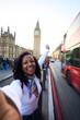 © CarlosBarquero - Vertical. Smiling african millennial woman taking a selfie in London with a red double-decker bus and Big Ben in the background. Travel portrait that conveys joy, sightseeing and city energy.