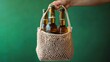 © btiger - Hand holding a reusable mesh bag with three brown beer bottles with green caps against a green background, relaxed casual eco-friendly mood