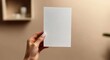 © zhuan - manicured hand holding blank white card against warm beige wall with blurred shelf and plant, calm minimalist mood