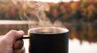© Rahmat - Cozy autumn lifestyle showing a close up of a hand holding a steaming black mug by a peaceful lake with colorful fall foliage reflections