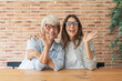 © Daniel - Mother and adult daughter wearing glasses sit at a wooden table, smiling and waving together in a casual home setting. Warm intergenerational bond, friendly greeting and relaxed lifestyle moment.