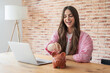 © Daniel - Happy young woman smiling and inserting coins into a piggy bank, symbolizing financial planning, savings, and investment goals while sitting at a wooden desk with a laptop