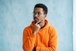© twilight mist - Thoughtful Young Black Man in Orange Hoodie and Glasses Looking Away Against Blue Background