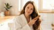 © Masque - A young woman brushes her long wavy brown hair with a wooden comb in the bathroom during the morning light.