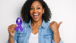© Luisa - Woman smiles, holding purple ribbon. She gestures thumbs-up with other hand. White background highlights her joyful expression. Curly hair frames her bright, happy face. Denim jacket adds casual