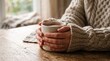 © Sadra - Person in cozy sweater holding a warm mug of coffee or tea at a wooden table