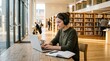 © Khan - Young woman studying with headphones at wooden table in bright modern library, typing on laptop with coffee cup, notebook, and shelves of books in background, education concept