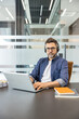 © Liubomir - Man with a headset and glasses looking at the camera while typing on a laptop, engaged in an online meeting or customer support role within a modern office setting