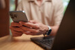 © eggeeggjiew - Asian man uses a smartphone beside a laptop at a desk, representing email communication, mobile productivity, online messaging, multitasking