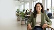 © Xenia - Smiling young businesswoman sitting at desk in modern office. Happy female professional in green blazer. Positive corporate workplace environment
