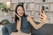 © Chadaporn - Young Asian woman taking selfie with smartphone at home, smiling and posing confidently while interacting with camera, representing social media activity, online