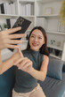 © Chadaporn - Young Asian woman taking selfie with smartphone at home, smiling and posing confidently while interacting with camera, representing social media activity, online