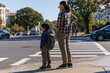 © Milos - A mother and her young son stand together at a crosswalk, showcasing a moment of connection and patience as they prepare to cross the street in a vibrant urban setting.