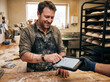 © FJ9 - Baker checking online orders on a tablet at a wooden table in his bakery, technology in a traditional food business