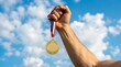 © Image - Hand of Victorious Champion Athlete Holding Up Gold Medal Against Blue Sky