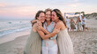 © Thumbs - Three women embracing on a beach at sunset with family in background