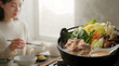 © Alongkorn - A young woman enjoys a comforting, steaming hot pot meal featuring fresh Asian vegetables and meats indoors