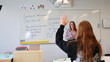 © Dark Artistic - Student raises her hand to ask a question while a teacher explains math problems on a whiteboard in a bright, modern classroom during an engaging lesson