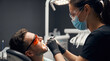 © Natali - A man sits in a dental chair at a clinic while a dental assistant performs an examination. The room has dental tools and bright lighting, and the man wears protective glasses