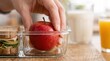 © Natali - A hand places a red apple into a glass lunch container alongside a sandwich. The setting is on a wooden surface with a glass of orange juice and a smoothie nearby