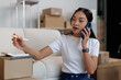 © David - Asian woman managing small business from home, multitasking with smartphone while calculating and preparing parcels for delivery, working on e commerce logistics