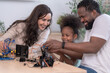 © ultramansk - An interracial family exploring STEM guide their child through a robotics kit at a bright learning table at home, sharing the joy of discovery captured in soft natural light.