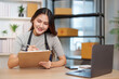 © Natee Meepian - Asian small business owner writing order notes on clipboard at home office desk