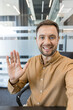 © Liubomir - Smiling man waving with an open hand, greeting during a virtual conference or online meeting from a modern office environment, engaging in digital communication