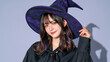© Maksym Tereshchenko - Cropped view of an Asian woman wearing a stylish purple witch hat and matching blouse with an elegant bow tie, face angled away from the camera, bright clean lavender background, modern Halloween of