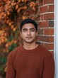 © CYBERPINK - Portrait of a young man standing in front of a brick wall. he is wearing a brown sweater and has a serious expression on his face.