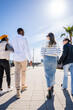 © CarlosBarquero - Vertical. Four diverse friends walk away together through a bright blue sky modern neighborhood, showing friendship, movement and open sunny outdoor lifestyle.
