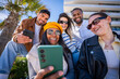 © CarlosBarquero - Multicultural friends sit closely together and smile while sharing content on a smartphone in a sunny modern residential district with palm tree and blue sky.