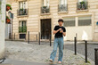 © dsbedarieva - A young man walks on a cobblestone street in Paris, France holding a plastic cup with iced coffee. He stands in front of classical residential buildings.