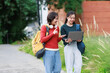 © Tj - Asian female students walking together on university campus, smiling and discussing coursework on a laptop, enjoying friendship and modern education outdoors