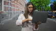 © Krakenimages.com - Woman with glasses holding laptop, typing with fingers on keyboard on street in front of brick building; concentration productivity remote work.