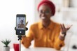© Prostock-studio - Smiling Black Female Content Creator Sitting At Home, Engaging In Live Video Presentation With Smartphone On Tripod, Creating Content For Social Media In A Relaxed Atmosphere, Business Concept.