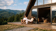 © lam - Description: A relaxed couple sit on wooden lounge chairs on the large terrace of a modern A‑frame cabin, admiring lush forest and rugged mountain scenery on a bright sunny day.