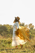 © Светлана  - A young woman strolls calmly and casually through a field; she is wearing an open yellow shawl and a brown hat, with her arms spread wide. Boho style.