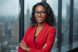 © polack - Confident black businesswoman smiles warmly in modern office space. She wears red blazer and stylish glasses. Her arms are crossed, posture exudes professionalism