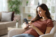 © PheelingsMedia - Woman sitting on armchair using cell phone at home