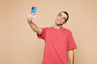 © ViDi Studio - Young middle eastern man wear red t-shirt casual clothes doing selfie shot on mobile cell phone post photo on social network isolated on plain pastel light beige background studio. Lifestyle concept.