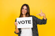 © Daniel - Woman smiling and pointing to a white sign reading vote in bold black letters, promoting civic participation, voting rights and democracy against a bright yellow background
