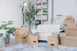 © Kritdanai - Moving Day Essentials: A contemporary living space is prepared for a move, with stacks of cardboard boxes, household items, and lush potted plants arranged on a plush rug, hinting at a fresh start.