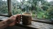 © MdShakil - Person holding a hot cup of coffee by a rainy window on a gloomy day
