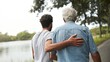 © Olya Pilipchik  - Adult man with arm around senior father's back, offering support and care during an outdoor walk together by the lake, showing intergenerational bond and assisting elderly parent
