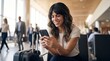 © Eve - Smiling female passenger using mobile payment application online banking app on smartphone holding phone sitting with luggage suitcase in airport waiting area prior to business departure